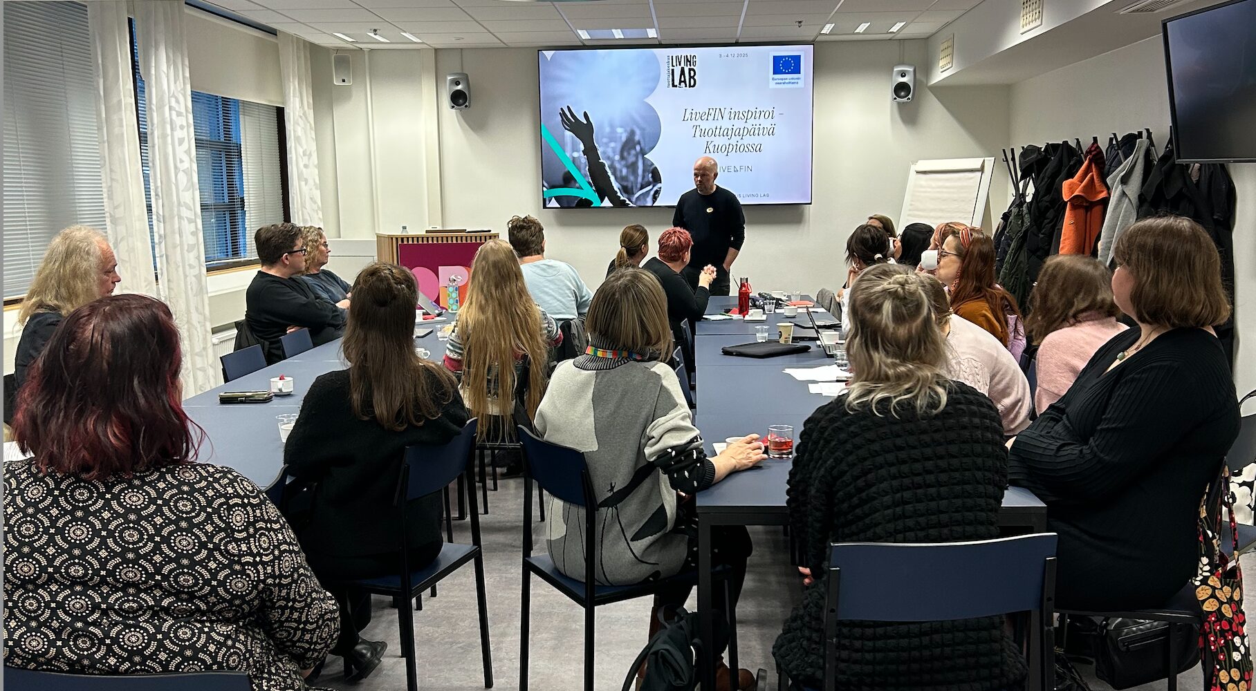 Joukko ihmisiä kuuntelee esittelyä huoneessa. Henkilö puhuu edessä. Esityksessä teksti: LivefIN inspiroi -tuottapäivät Kuopiossa.
